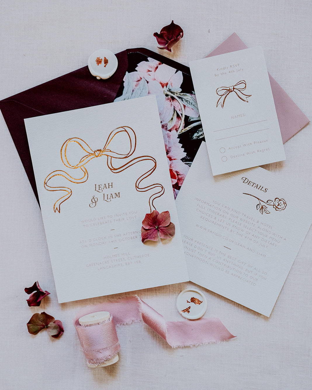 Wedding Invitation featuring a large Rose Gold Bow Design, foiled onto thick textured card with couples names and information printed in a soft brown colour. Paired with burgundy envelope with floral envelope liner, white wax seal with rose gold flakes and blush pink silk ribbon. Laid out flat lay style with Details card and RSVP. Designed by Fancy Folk Wedding Stationery