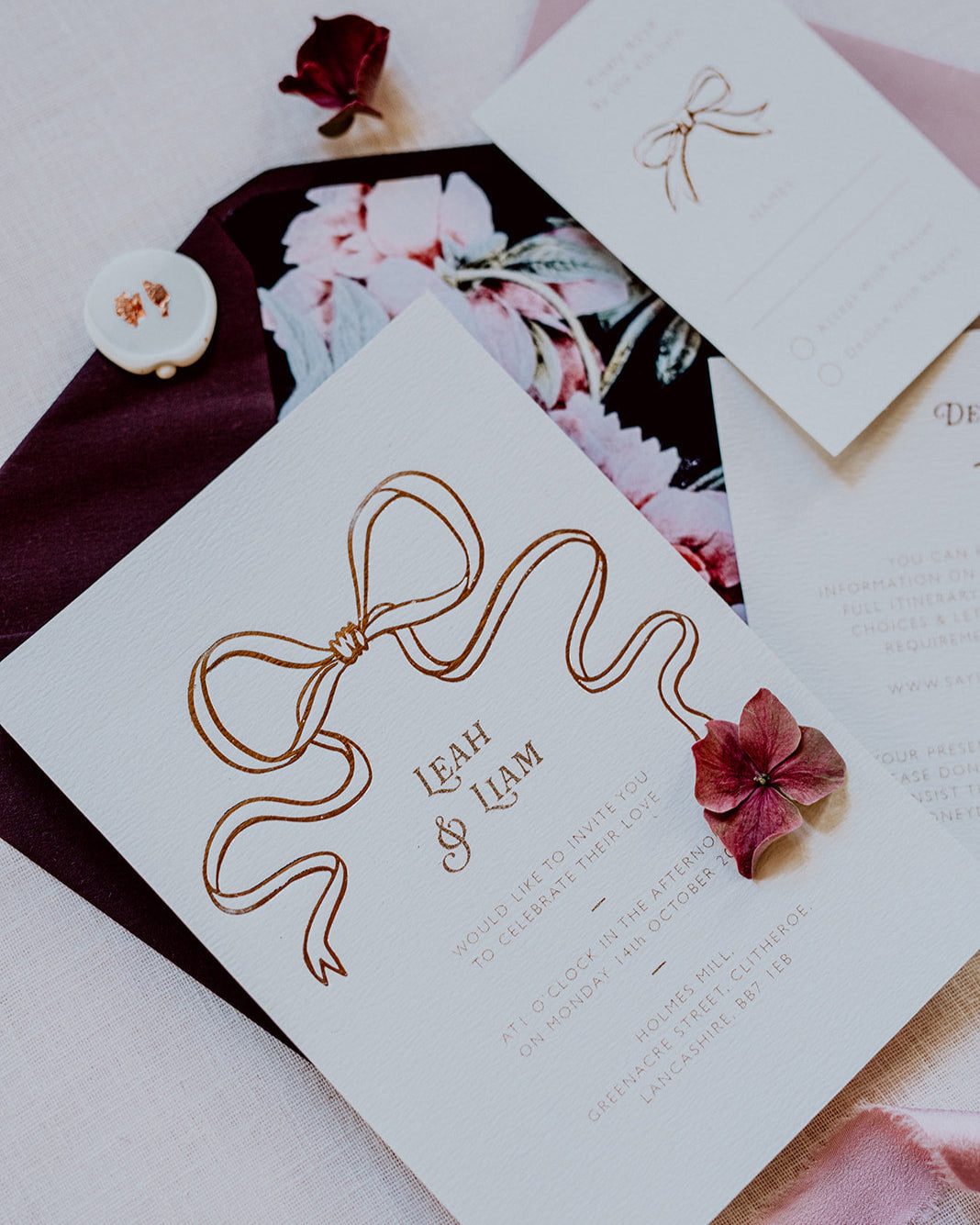 Close up of a wedding Invitation featuring a large Rose Gold Bow Design, foiled onto thick textured card with couples names and information printed in a soft brown colour. Paired with burgundy envelope with floral envelope liner, white wax seal with rose gold flakes and blush pink silk ribbon. Designed by Fancy Folk Wedding Stationery