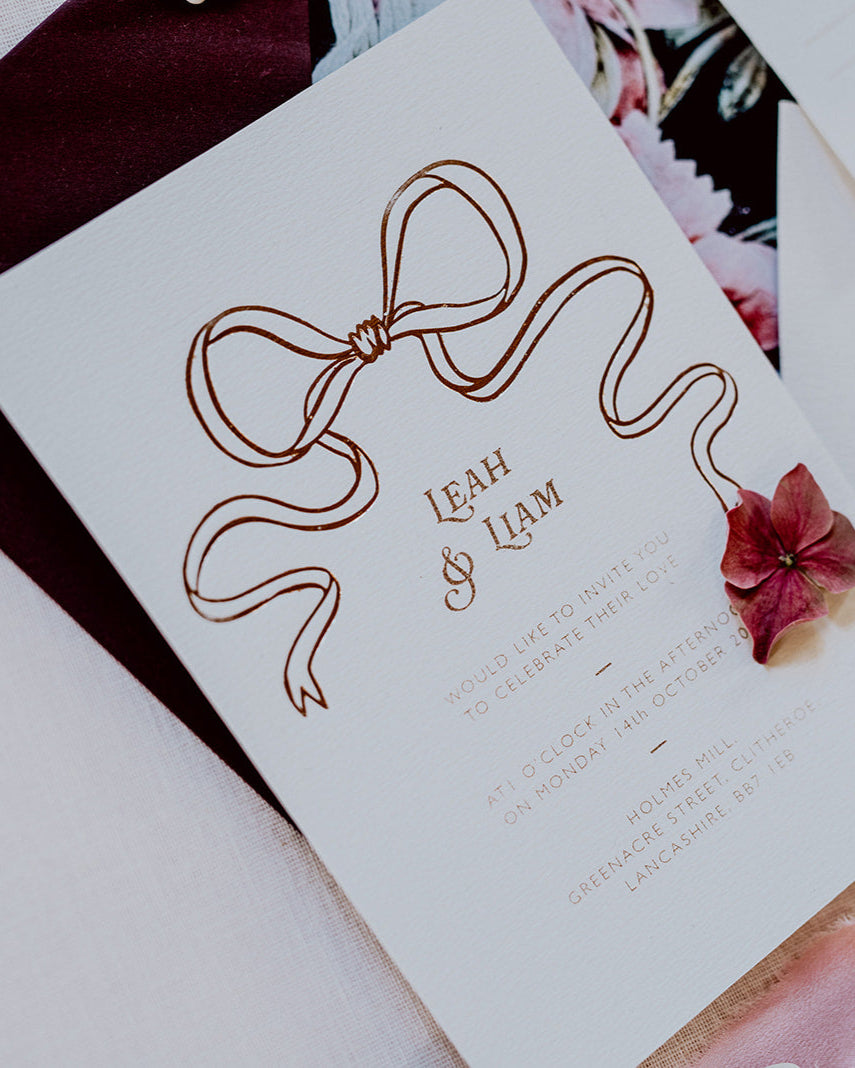 Close up of a wedding Invitation featuring a large Rose Gold Bow Design, foiled onto thick textured card with couples names and information printed in a soft brown colour. Paired with burgundy envelope with floral envelope liner, white wax seal with rose gold flakes and blush pink silk ribbon. Designed by Fancy Folk Wedding Stationery