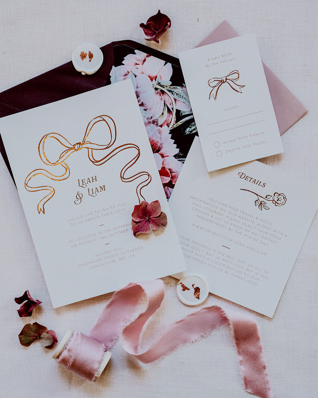 Wedding Invitation featuring a large Rose Gold Bow Design, foiled onto thick textured card with couples names and information printed in a soft brown colour. Paired with burgundy envelope with floral envelope liner, white wax seal with rose gold flakes and blush pink silk ribbon. Laid out flat lay style with Details card and RSVP. Designed by Fancy Folk Wedding Stationery