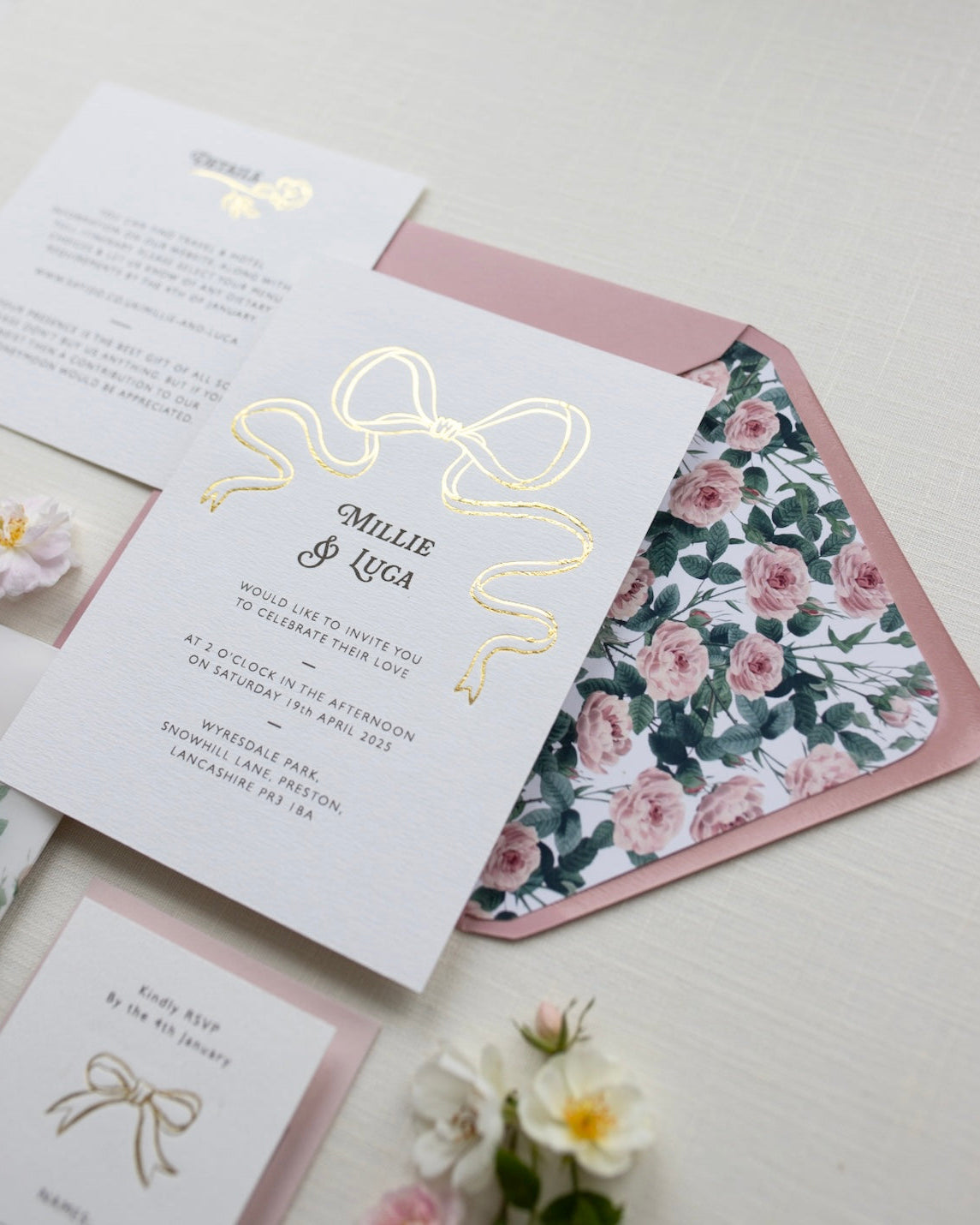 Bow Wedding Invitation featuring a large Gold Bow Design, foiled onto thick textured card with couples names and information printed in a soft brown colour. Paired with dusky pink envelope with english rose garden floral envelope liner. Laid out flat lay style with Details card and RSVP and wild roses. Designed by Fancy Folk Wedding Stationery