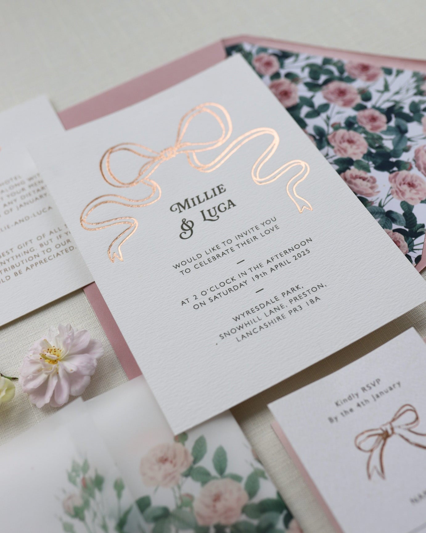 Bow Wedding Invitation featuring a large Gold Bow Design, foiled onto thick textured card with couples names and information printed in a soft brown colour. Paired with dusky pink envelope with english rose garden floral envelope liner. Laid out flat lay style with Details card and RSVP and wild roses. Designed by Fancy Folk Wedding Stationery
