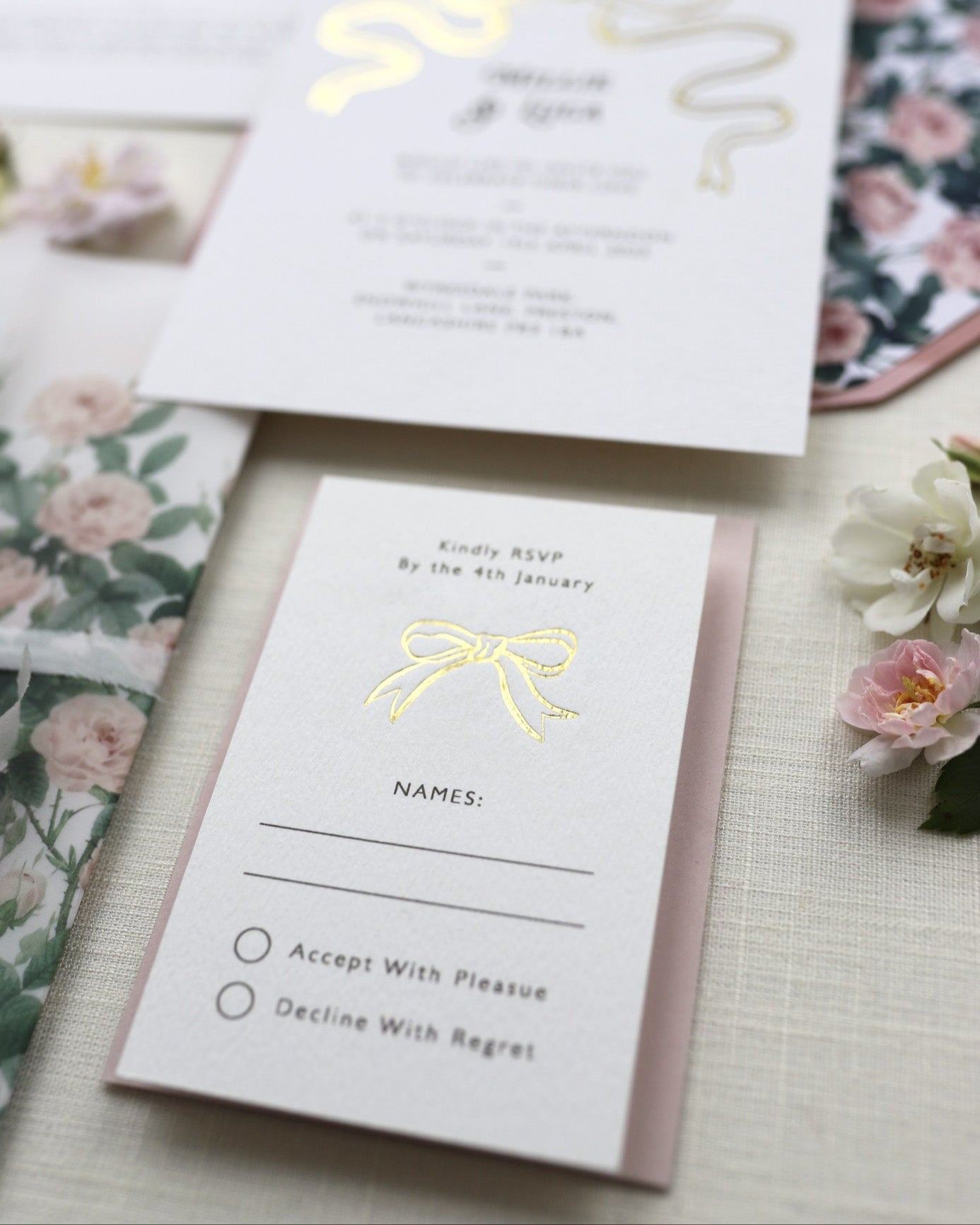Bow Wedding RSVP featuring a small Gold Bow Design, foiled onto thick textured card with couples names and information printed in a soft brown colour. Paired with dusky pink envelope. Designed by Fancy Folk Wedding Stationery