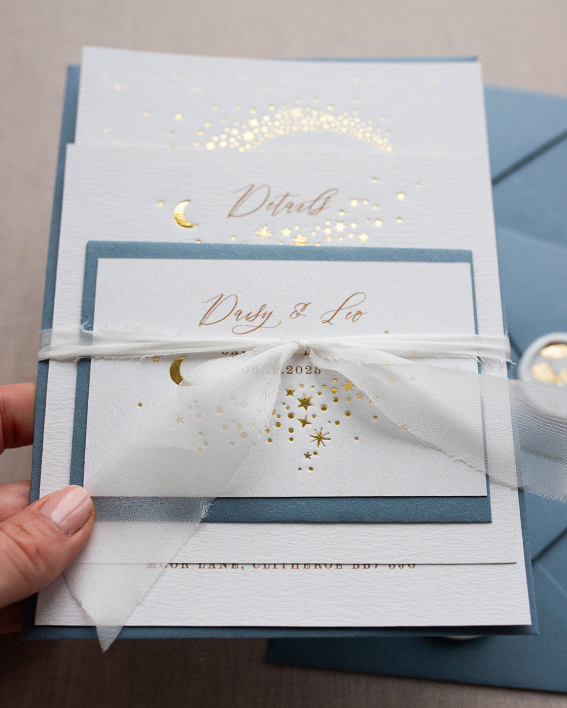 Celestial wedding stationery sample pack. Invitation,Details card and RSVP layer on top of each other, tied together in a perfect bundle with white silk ribbon. A truly magical crescent moon design made up of lots of little stars is hot foiled in gold on to the main invitation. Smaller cluster of gold stars feature on the details card and RSVP. The invitations are foiled and printed onto thick textured card, styled with silk ribbon, wax seals and dusty blue coloured envelopes. FancyFolk Design