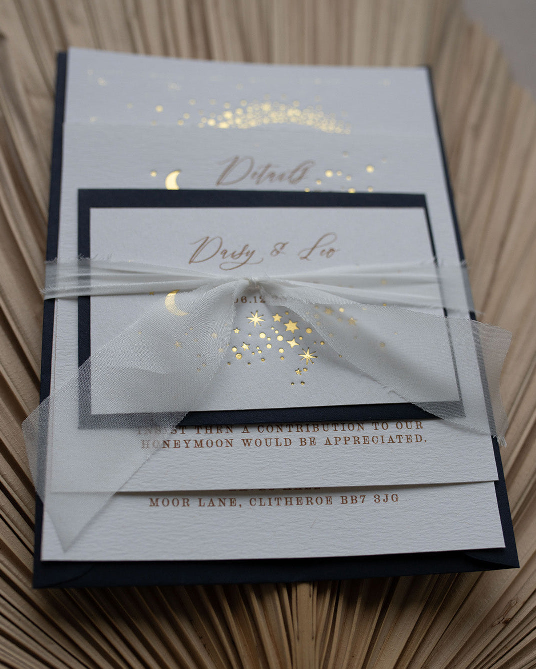 Celestial wedding stationery sample pack. Invitation,Details card and RSVP layer on top of each other, tied together in a perfect bundle with white silk ribbon. A truly magical crescent moon design made up of lots of little stars is hot foiled in gold on to the main invitation. Smaller cluster of gold stars feature on the details card and RSVP. The invitations are foiled and printed onto thick textured card, styled with silk ribbon, wax seals and navy blue coloured envelopes. FancyFolk Design