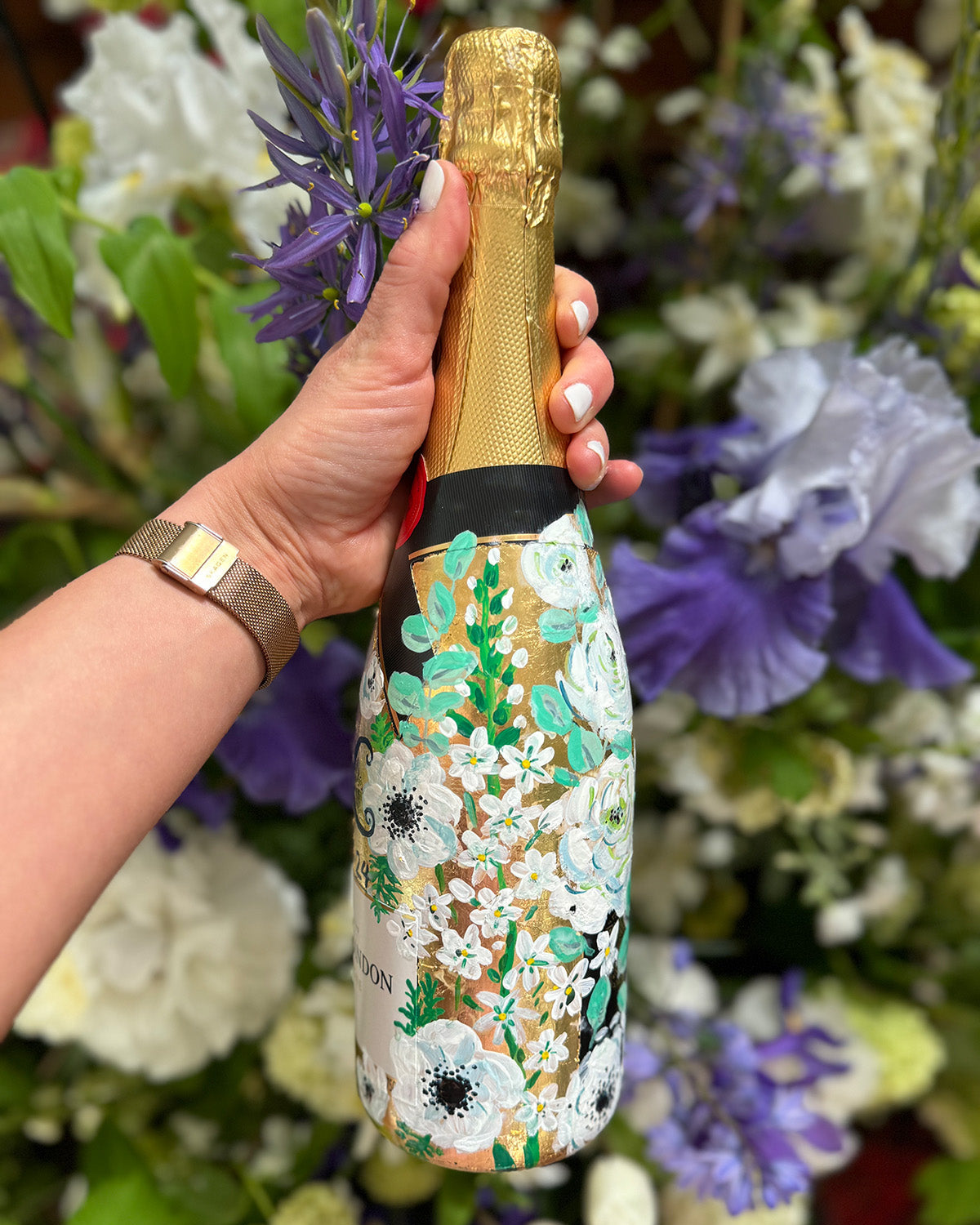 Hand painted Bottle of Moet Champagne. Gold leaf base painted florals to match the bridal bouquet flowers. Woman's hands holding the bottle. Fancy Folk Design