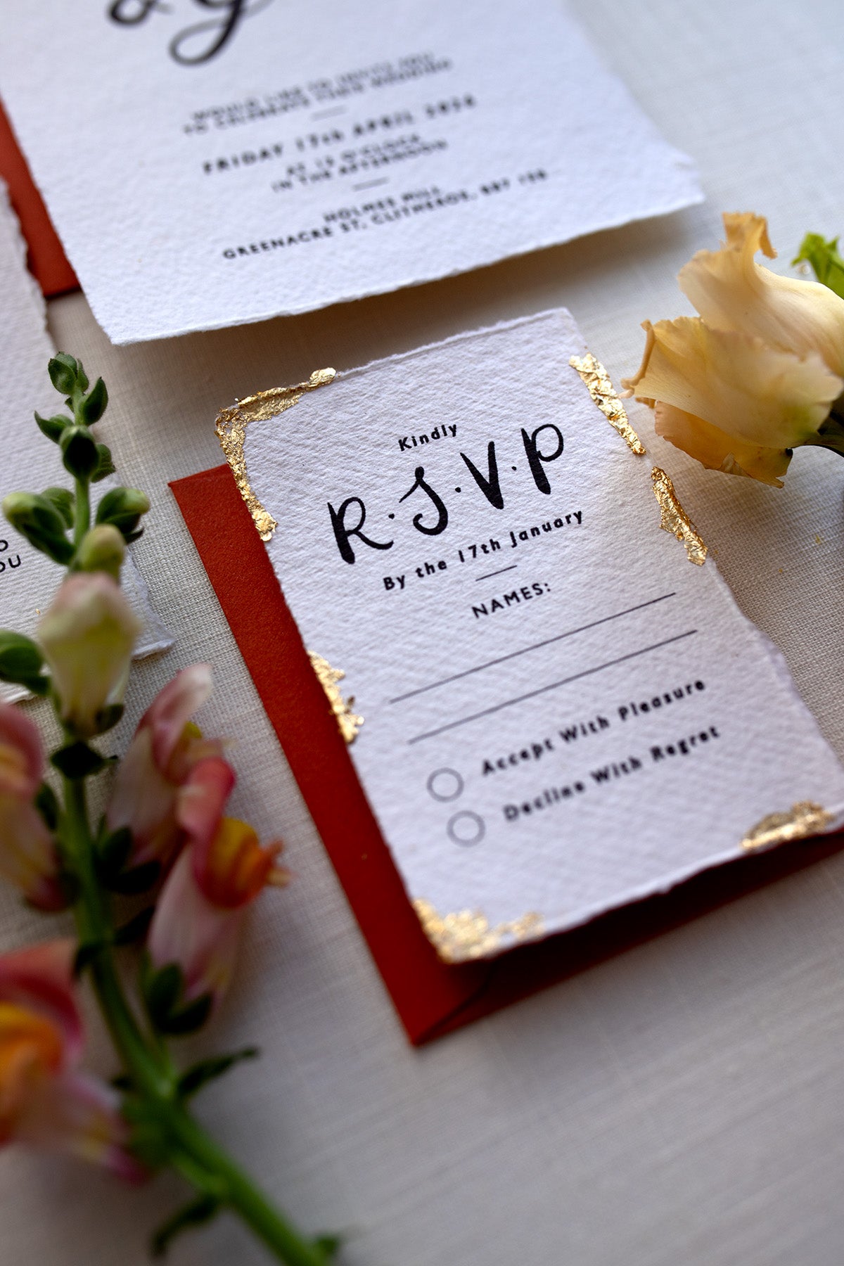Modern Calligraphy Wedding RSVP card with terracotta coloured envelopes. Our wedding stationery features a beautiful hand lettered calligraphy names design printed on textured handmade Indian paper with hand torn edges for a soft romantic feel. Each piece is lovingly hand finished with gold leaf for a luxury feel. Styled with white and gold leaf wax seals and peachy florals. Fancy Folk