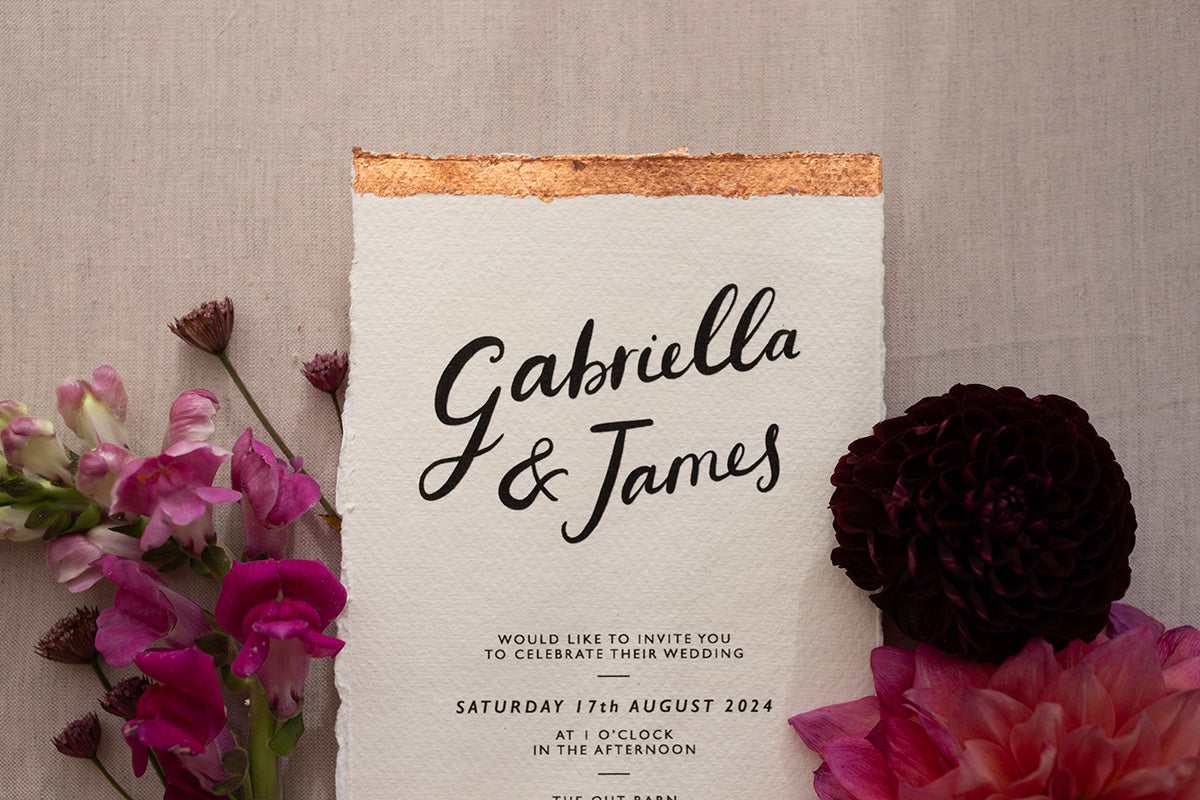 Close up of Modern Calligraphy Wedding Invitation, with rich burgundy coloured envelopes.Our wedding stationery features a beautiful hand lettered calligraphy names design printed on textured handmade Indian paper with hand torn edges for a soft romantic feel. Each piece is lovingly hand finished with copper leaf for a luxury feel. Styled with burgundy and coral florals, perfect for autumn and winter weddings. Fancy Folk