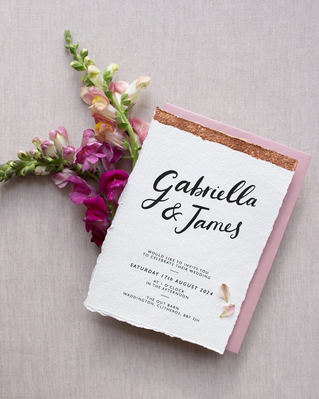 Modern Calligraphy Wedding Invitation, with blush pink coloured envelopes. Our wedding stationery features a beautiful hand lettered calligraphy names design printed on textured handmade Indian paper, with hand torn edges for a soft romantic feel. Each piece is lovingly hand finished with copper leaf for a luxury feel. Styled with burgundy and pink florals. Fancy Folk
