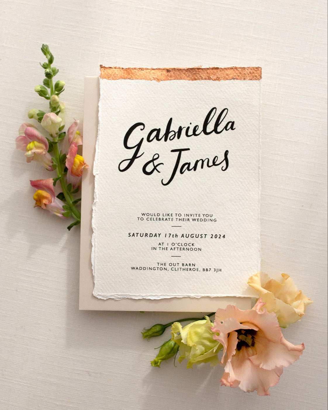 Modern Calligraphy Wedding Invitation, with neutral almond coloured envelope. Features a beautiful hand lettered calligraphy names design printed on textured handmade Indian paper with hand torn edges for a soft romantic feel. Each piece is lovingly hand finished with copper leaf for a luxury feel. Styled with peachy florals. Fancy Folk