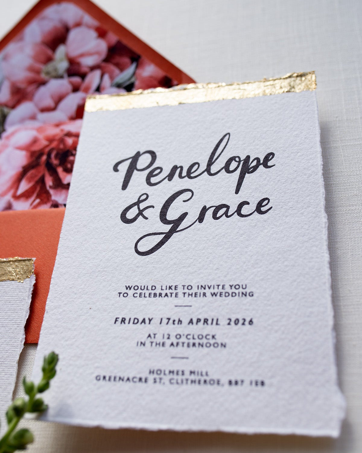 A close up of our Modern Calligraphy Wedding Invitation with terracotta coloured envelopes. Our wedding stationery features a beautiful hand lettered calligraphy names design printed on textured handmade Indian paper with hand torn edges for a soft romantic feel. Each piece is lovingly hand finished with gold leaf for a luxury feel. Styled with white and gold leaf wax seals and peachy florals. Fancy Folk