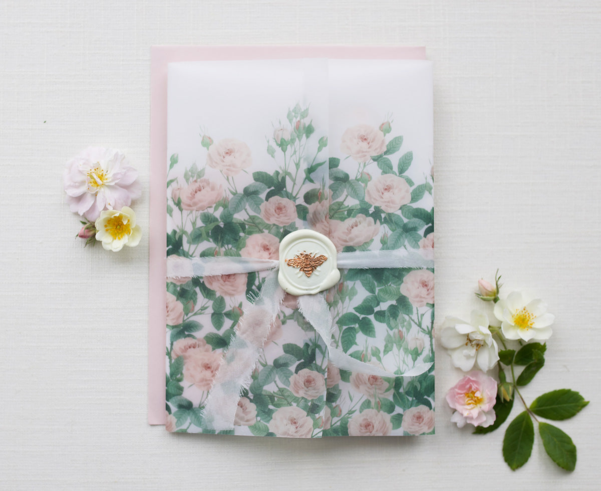 A wedding Stationery Suite wrapped in a rose garden design vellum wrap, tied with a white Italian silk ribbon, finished with a white and rose gold bee wax seal. Styles with wild roses. Fancy Folk Wedding Stationery