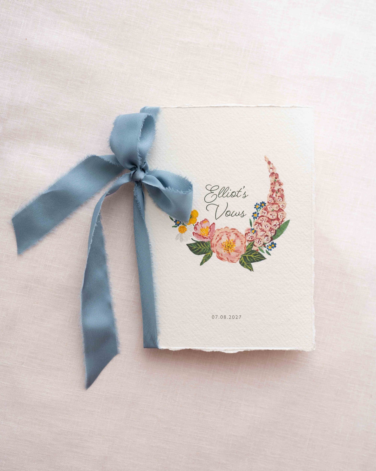 A wild flower vow book. Watercolour hand painted florals of meadow flowers and a little bee make up a posy. The words Elliots Vows are nestled in the floral with wedding date set below. Printed on handmade Indian cotton paper and styled with dusky blue silk ribbon. With a blush pink background. Fancy Folk Design