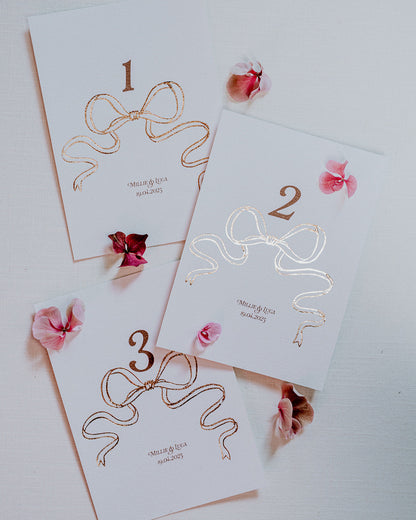 Rose Gold Foil Bow design Wedding Table Numbers with printed couples names and wedding date. Designed by Fancy Folk Wedding Sationery.