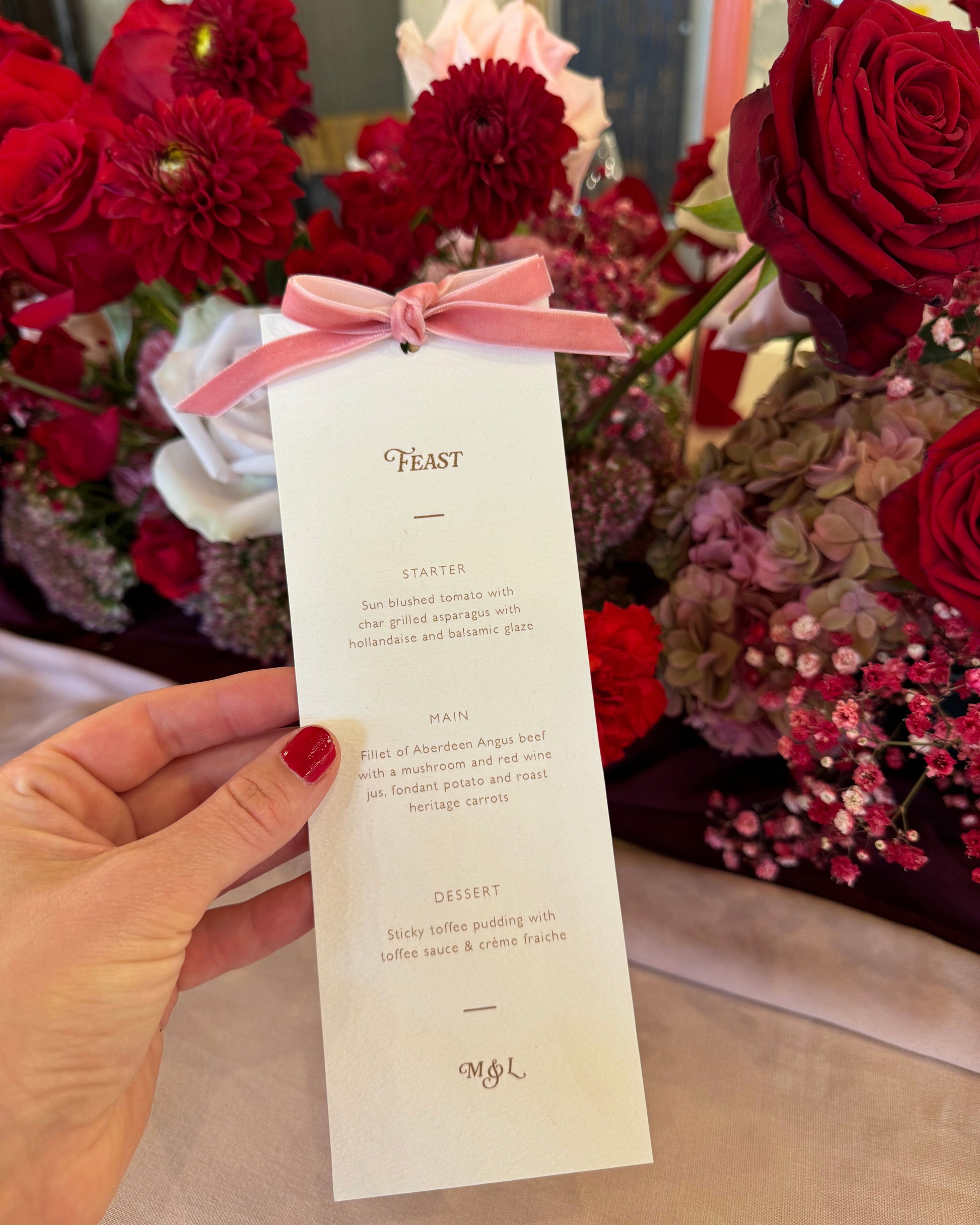 Hand holding a blush velvet bow wedding breakfast menu, with red floral arrnagment behind. Designed by Fancy Folk Wedding Stationery