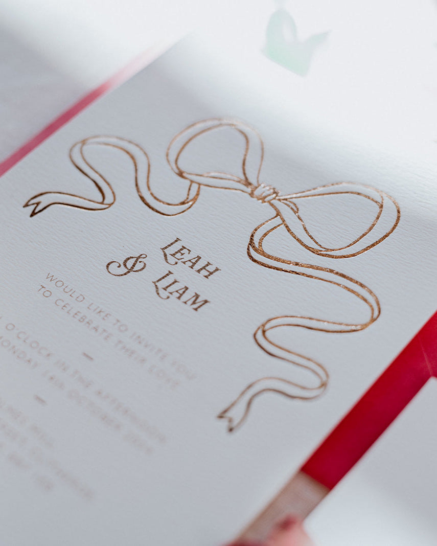 Close up of a wedding Invitation featuring a large Rose Gold Bow Design, foiled onto  thick textured card with couples names and information printed in a soft brown colour. Designed by Fancy Folk Wedding Stationery