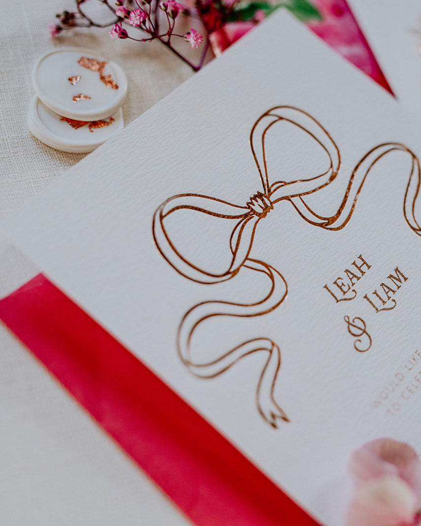 Close up of a wedding Invitation featuring a large Rose Gold Bow Design, foiled onto  thick textured card with couples names and information printed in a soft brown colour. Paired with hot pink envelope and white wax seal with rose gold flakes. Designed by Fancy Folk Wedding Stationery