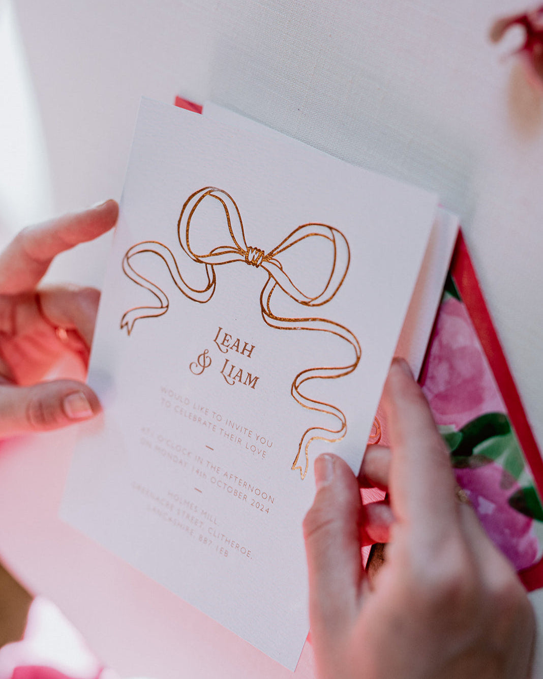 Close up of a wedding Invitation featuring a large Rose Gold Bow Design, foiled onto  thick textured card with couples names and information printed in a soft brown colour. Held in a ladies hands as she opens the invitation. Paired with hot pink envelope with floral envelope liner, white wax seal with rose gold flakes. Designed by Fancy Folk Wedding Stationery