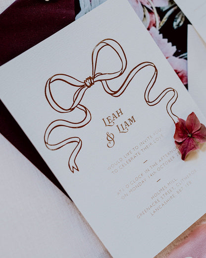 Close up of a wedding Invitation featuring a large Rose Gold Bow Design, foiled onto  thick textured card with couples names and information printed in a soft brown colour. Paired with burgundy envelope with floral envelope liner, white wax seal with rose gold flakes and blush pink silk ribbon. Designed by Fancy Folk Wedding Stationery