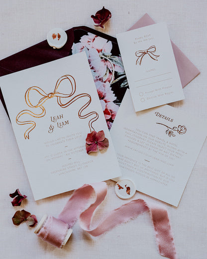 Wedding Invitation featuring a large Rose Gold Bow Design, foiled onto  thick textured card with couples names and information printed in a soft brown colour. Paired with burgundy envelope with floral envelope liner, white wax seal with rose gold flakes and blush pink silk ribbon. Laid out flat lay style with Details card and RSVP. Designed by Fancy Folk Wedding Stationery