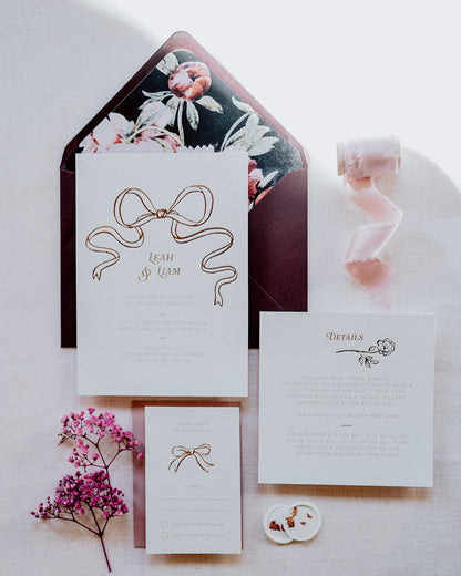 Wedding Invitation Sample Pack featuring a large Rose Gold Bow Design, foiled onto  thick textured card with couples names and information printed in a soft brown colour. Paired with burgundy envelope with floral envelope liner, white wax seal with rose gold flakes and blush pink silk ribbon. Laid out flat lay style with Details card and RSVP. Designed by Fancy Folk Wedding Stationery