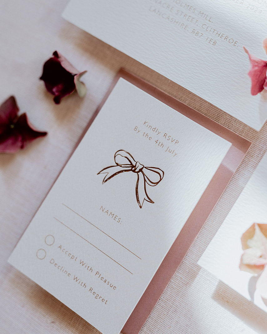 Close up of wedding RSVP card featuring a small foiled bow design below the word 'Kindly RSVP'. Foiled onto thick textured card with couples  information printed in a soft brown colour. Paired a dusty pink envelope. Designed by Fancy Folk Wedding Stationery