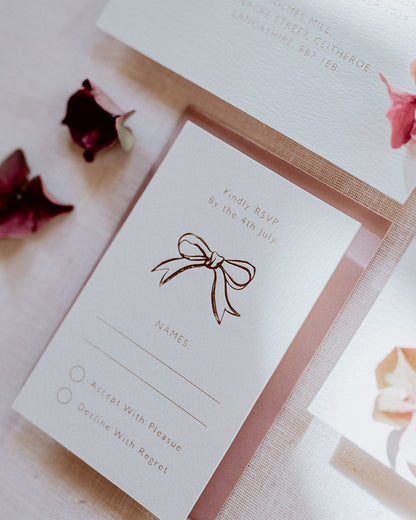 Close up of wedding RSVP card featuring a small foiled bow design below the word 'Kindly RSVP'. Foiled onto thick textured card with couples  information printed in a soft brown colour. Paired a dusty pink envelope. Designed by Fancy Folk Wedding Stationery
