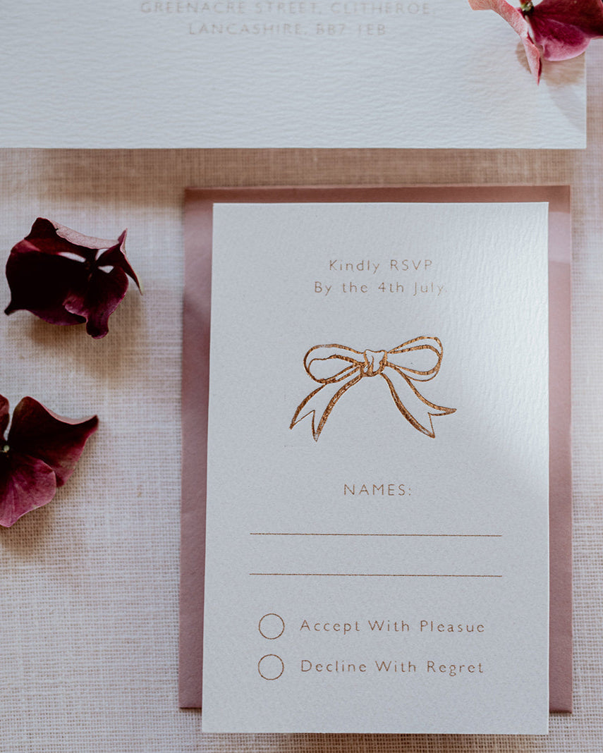 Close up of wedding RSVP card featuring a small foiled bow design below the word 'Kindly RSVP'. Foiled onto thick textured card with couples  information printed in a soft brown colour. Paired a dusty pink envelope. Designed by Fancy Folk Wedding Stationery