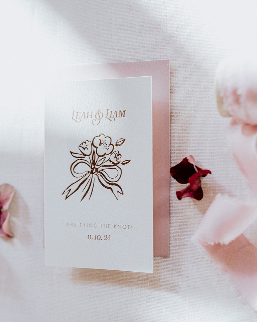 Bow Posy Wedding Save The Date Card with Dusty Pink envelope. Hot foil flower and bow icon printed onto thick GFSmith Card with couples names and wedding date. "Leah & Liam are tying the knot! 11.10.24" Designed by Fancy Folk Wedding Stationery.
