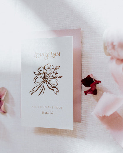 Bow Posy Wedding Save The Date Card with Dusty Pink envelope. Hot foil flower and bow icon printed onto thick GFSmith Card with couples names and wedding date. "Leah & Liam are tying the knot! 11.10.24" Designed by Fancy Folk Wedding Stationery.