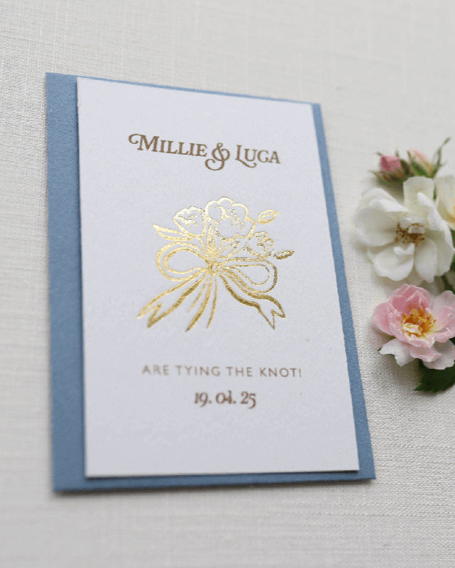 Bow Posy Wedding Save The Date Card with Dusty Blue envelope. Gold Hot foil printed onto thick GFSmith Card with couples names and wedding date. "Millie & Luca are tying the knot! 19.04.25" Designed by Fancy Folk Wedding Stationery. 