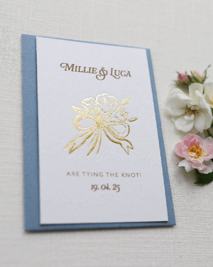 Bow Posy Wedding Save The Date Card with Dusty Blue envelope. Gold Hot foil printed onto thick GFSmith Card with couples names and wedding date. "Millie & Luca are tying the knot! 19.04.25" Designed by Fancy Folk Wedding Stationery. 