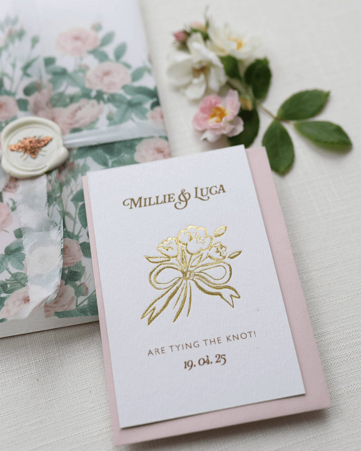Bow Posy Wedding Save The Date Card with Dusty Pink envelope. Gold Hot foil printed onto thick GFSmith Card with couples names and wedding date. "Millie & Luca are tying the knot! 19.04.25" Designed by Fancy Folk Wedding Stationery. 