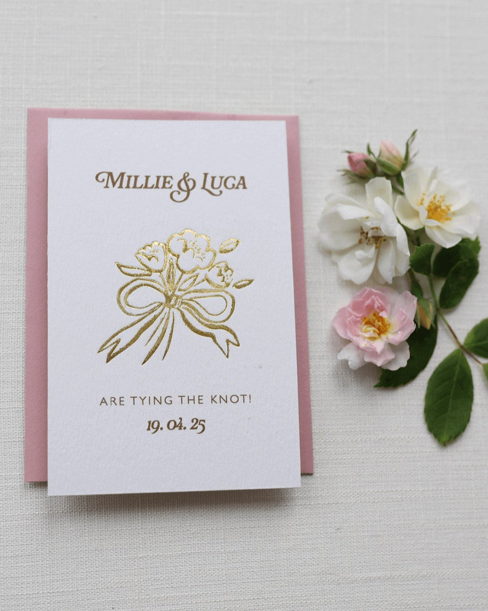 Bow Posy Wedding Save The Date Card with Dusty Pink envelope. Gold Hot foil printed onto thick GFSmith Card with couples names and wedding date. "Millie & Luca are tying the knot! 19.04.25" Designed by Fancy Folk Wedding Stationery. 