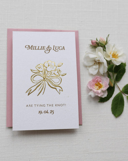 Bow Posy Wedding Save The Date Card with Dusty Pink envelope. Gold Hot foil printed onto thick GFSmith Card with couples names and wedding date. "Millie & Luca are tying the knot! 19.04.25" Designed by Fancy Folk Wedding Stationery. 
