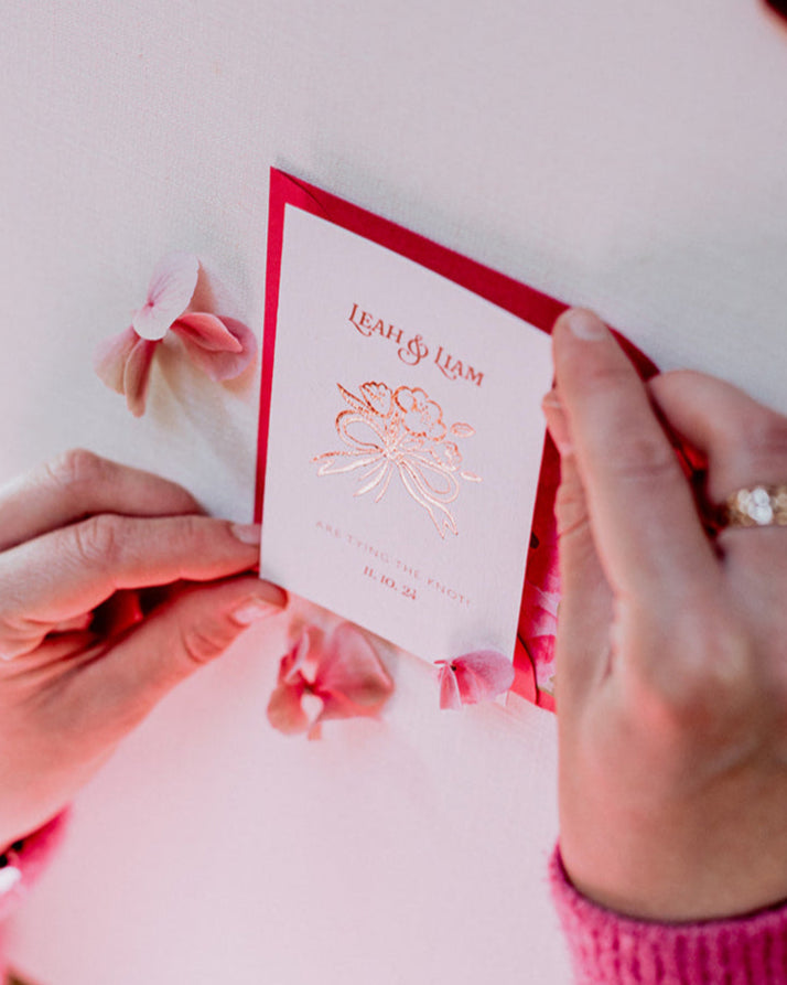 Bow Posy Wedding Save The Date Card with Hot Pink floral envelope liners. Hot foil printed onto thick GFSmith Card with couples names and wedding date. "Leah & Liam are tying the knot! 11.10.24" Designed by Fancy Folk Wedding Stationery. Stylist setting up stationery for a photoshoot.