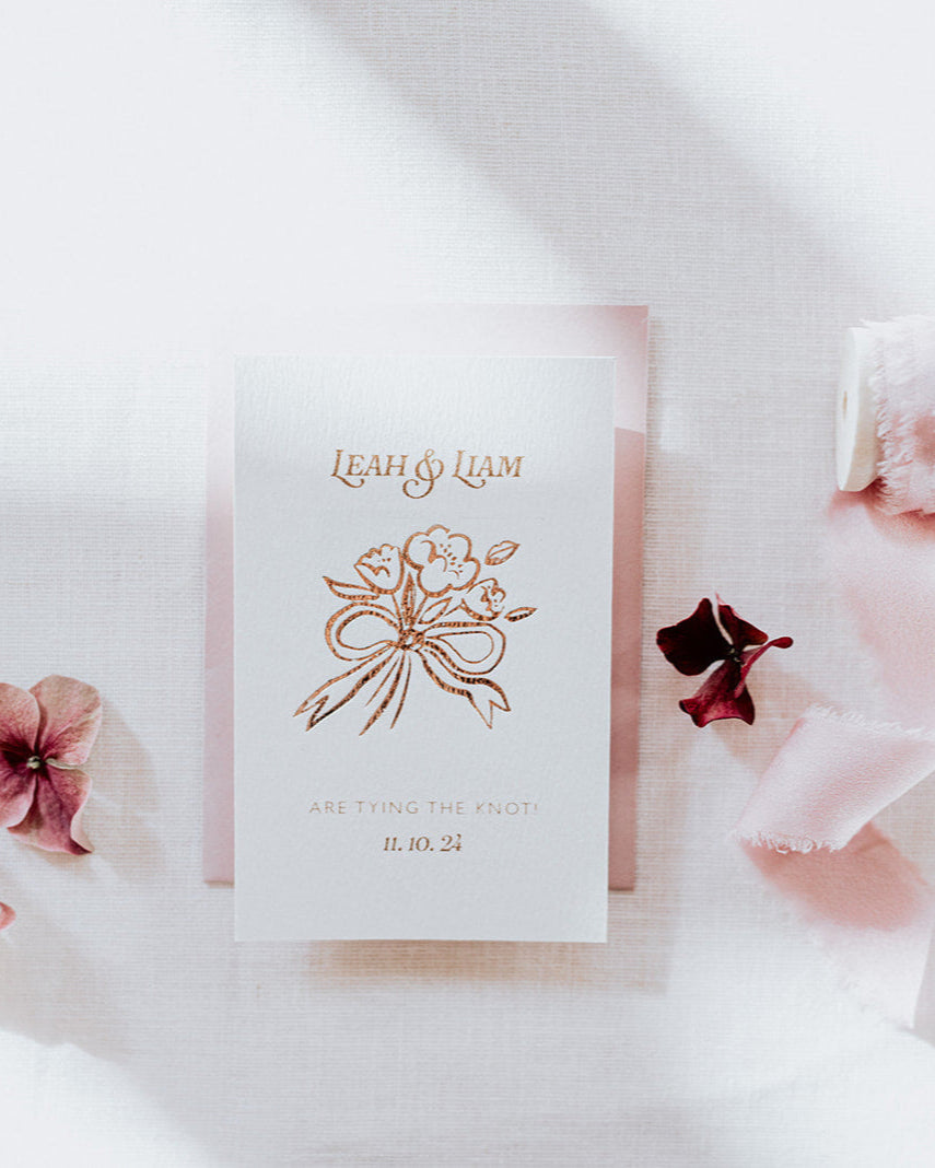 Bow Posy Wedding Save The Date Card with Dusty Pink envelope. Hot foil printed onto thick GFSmith Card with couples names and wedding date. "Leah & Liam are tying the knot! 11.10.24" Designed by Fancy Folk Wedding Stationery. 