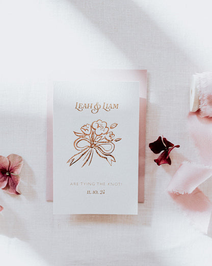 Bow Posy Wedding Save The Date Card with Dusty Pink envelope. Hot foil printed onto thick GFSmith Card with couples names and wedding date. "Leah & Liam are tying the knot! 11.10.24" Designed by Fancy Folk Wedding Stationery. 