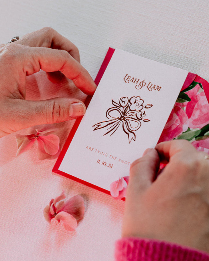 Bow Posy Wedding Save The Date Card with Hot Pink floral envelope liners. Hot foil printed onto thick GFSmith Card with couples names and wedding date. "Leah & Liam are tying the knot! 11.10.24" Designed by Fancy Folk Wedding Stationery. Close up of hand holding the crad.