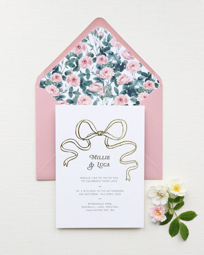 Bow Wedding Invitation featuring a large Gold Bow Design, foiled onto  thick textured card with couples names and information printed in a soft brown colour. Paired with dusky pink envelope with english rose garden floral envelope liner. Designed by Fancy Folk Wedding Stationery