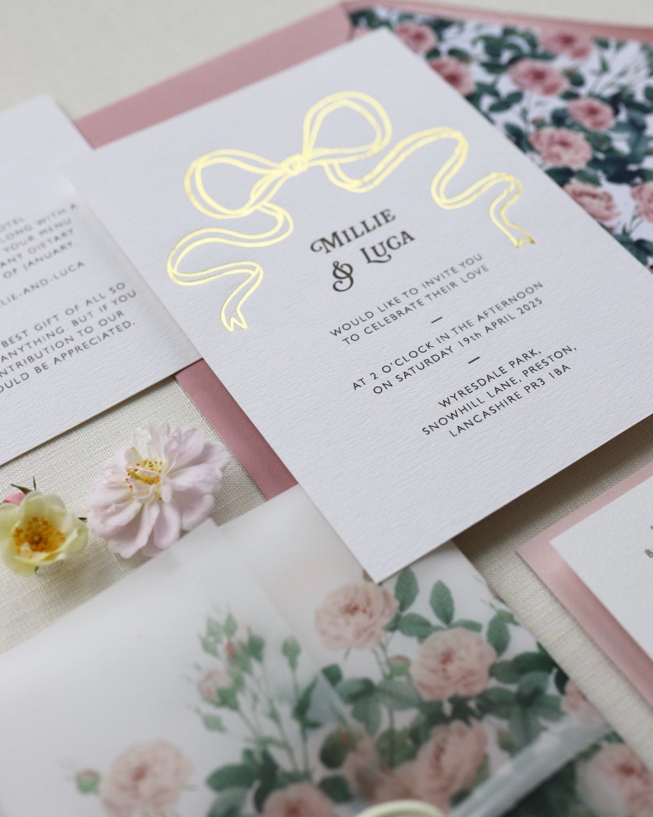 Close up of Bow Wedding Invitation featuring a large Gold Bow Design, foiled onto  thick textured card with couples names and information printed in a soft brown colour. Paired with dusky pink envelope with english rose garden floral envelope liner, vellum wrap and wax seal. Laid out flat lay style with Details card and RSVP and wild roses. Designed by Fancy Folk Wedding Stationery