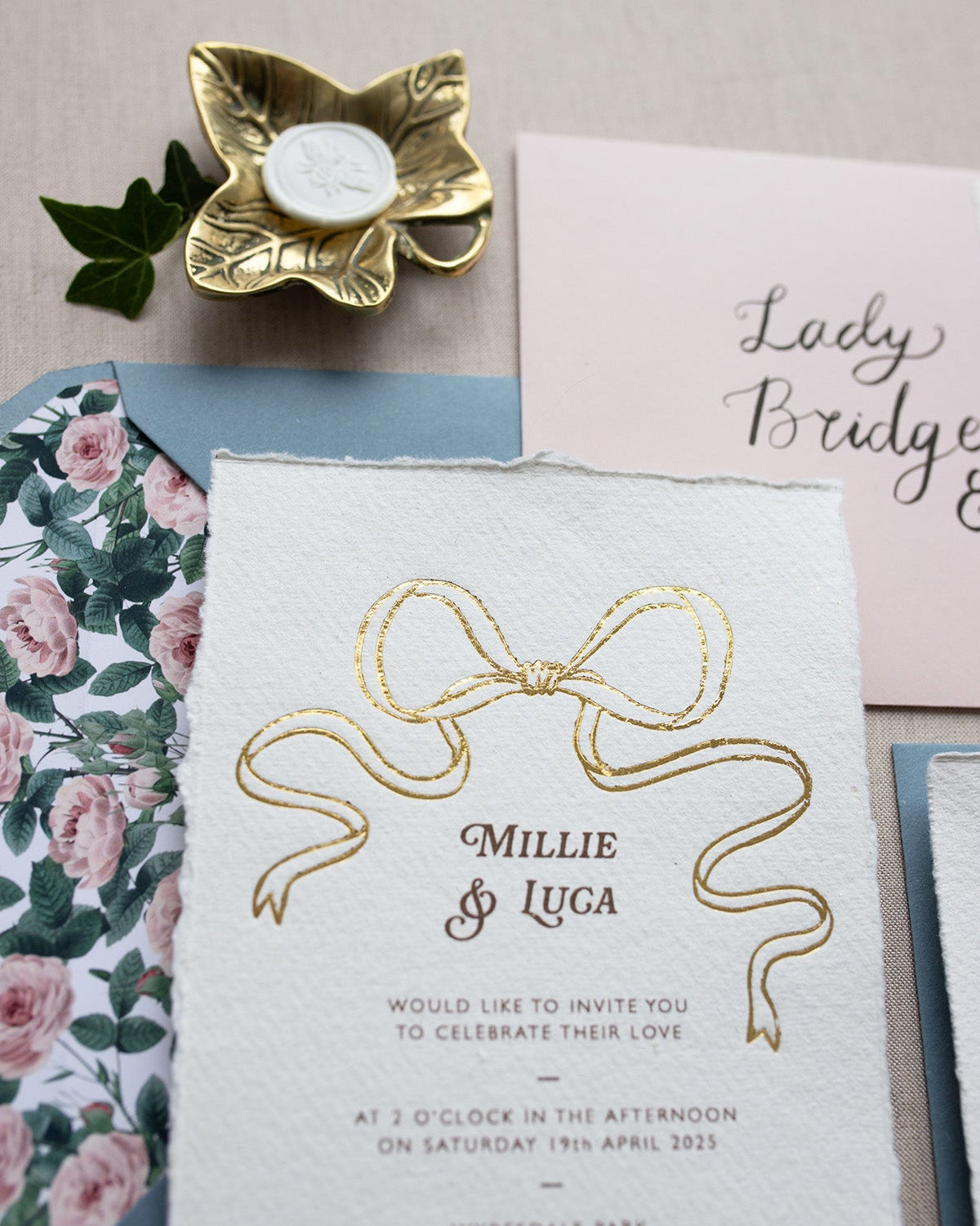 Close up of gold Bow detail wedding invitation. Paired with dusty blue coloured envelope and rose garden envelope liner. Printed on luxury hand made cotton paper with romantic hand torn edges. Styles with gold Ivy trinket dish and bee wax seal. Designed by Fancy Folk Weddings