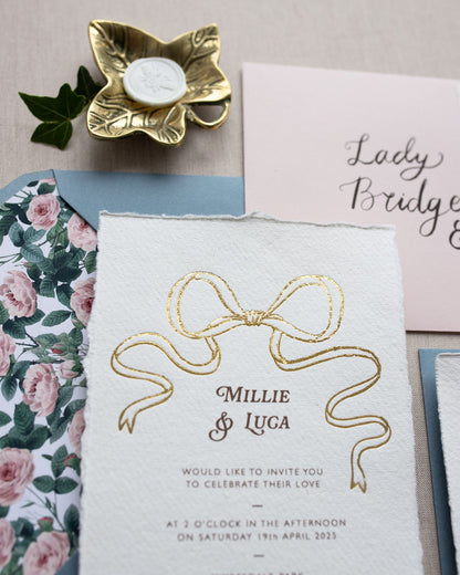 Close up of gold Bow detail wedding invitation. Paired with dusty blue coloured envelope and rose garden envelope liner. Printed on luxury hand made cotton paper with romantic hand torn edges. Styles with gold Ivy trinket dish and bee wax seal. Designed by Fancy Folk Weddings