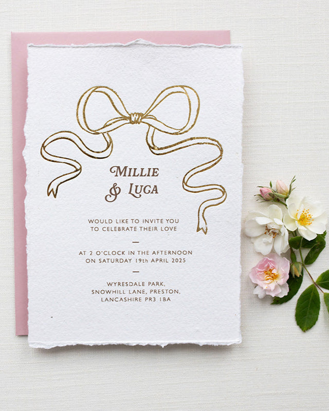 Gold Bow detail wedding invitation. Paired with blush pink coloured envelope. Printed on luxury hand made cotton paper with romantic hand torn edges. Styled with wild roses. Designed by Fancy Folk Weddings