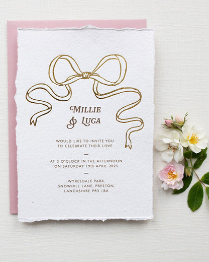 Gold Bow detail wedding invitation. Paired with blush pink coloured envelope. Printed on luxury hand made cotton paper with romantic hand torn edges. Styled with wild roses. Designed by Fancy Folk Weddings