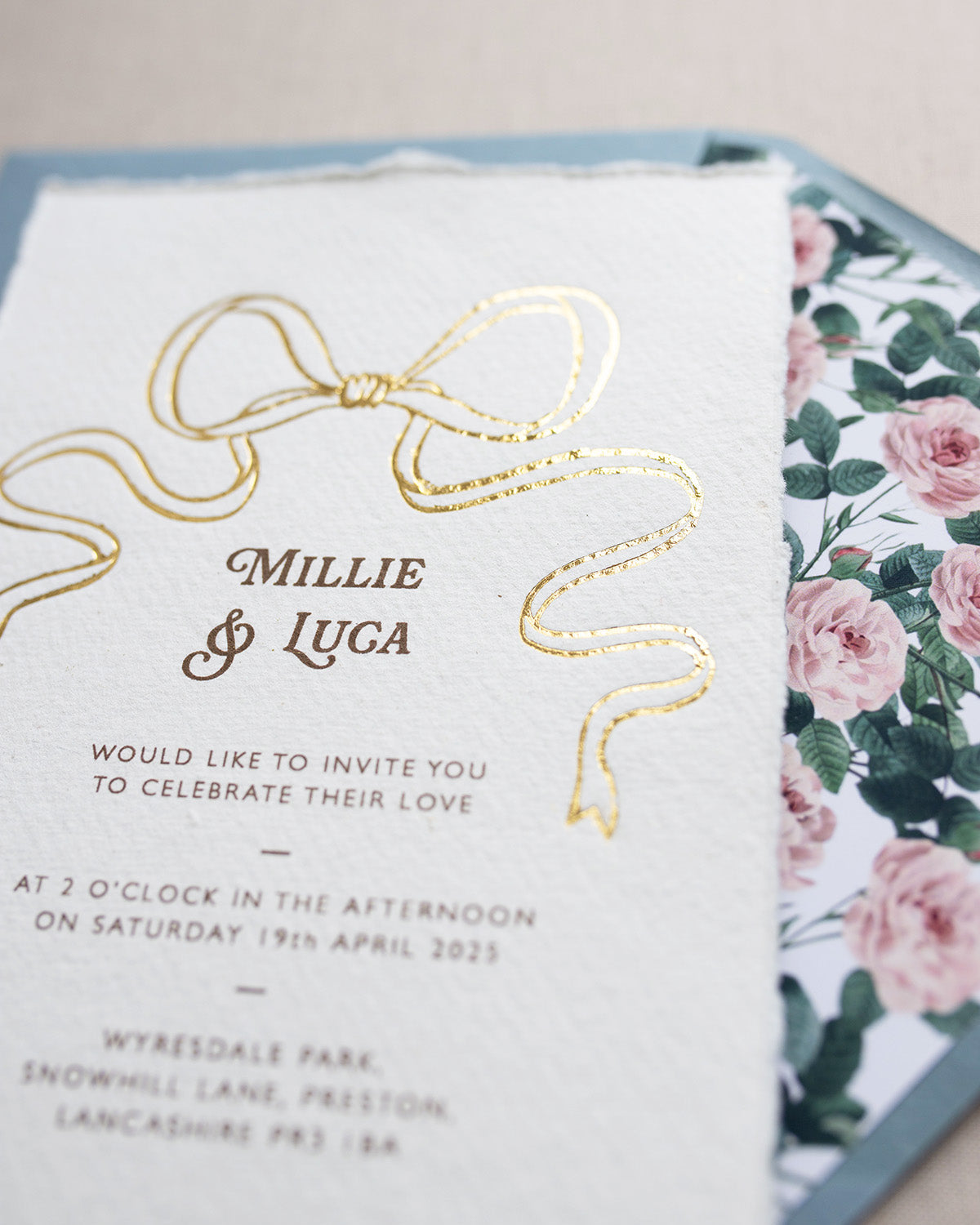 Close up of gold Bow detail wedding invitation. Paired with dusty blue coloured envelope and rose garden envelope liner. Printed on luxury hand made cotton paper with romantic hand torn edges. Designed by Fancy Folk Weddings