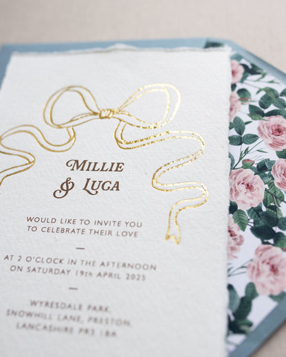Close up of gold Bow detail wedding invitation. Paired with dusty blue coloured envelope and rose garden envelope liner. Printed on luxury hand made cotton paper with romantic hand torn edges. Designed by Fancy Folk Weddings