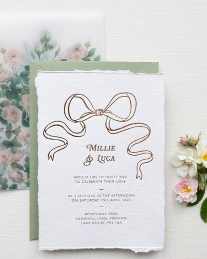 Bridgerton Inspired Rose Gold Foil Bow design wedding invitation. Sage green colour envelope paired with floral rose garden envelope liner and semi transparent rose print vellum wrap. Printed on a luxurious and tactile handmade cotton paper. Flat lay styled  with wild roses. Designed by Fancy Folk Weddings