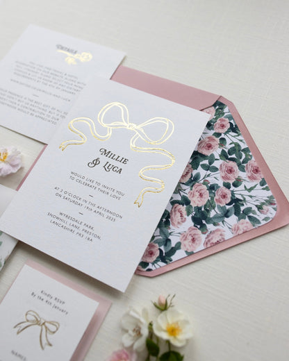 Bow Wedding Invitation featuring a large Gold Bow Design, foiled onto  thick textured card with couples names and information printed in a soft brown colour. Paired with dusky pink envelope with english rose garden floral envelope liner. Laid out flat lay style with Details card and RSVP and wild roses. Designed by Fancy Folk Wedding Stationery
