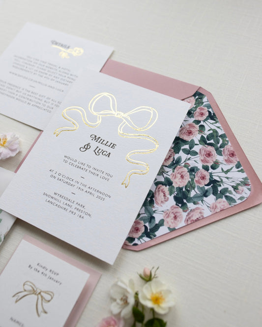 Bow Wedding Invitation featuring a large Gold Bow Design, foiled onto  thick textured card with couples names and information printed in a soft brown colour. Paired with dusky pink envelope with english rose garden floral envelope liner. Laid out flat lay style with Details card and RSVP and wild roses. Designed by Fancy Folk Wedding Stationery